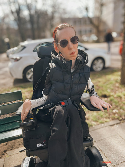 Ich sitze neben einer Parkbank und schaue nachdenklich in die Ferne. Ich trage ein asymmetrisches weiß graues Shirt, eine schwarze Weste, schwarze Jeans, sowie eine goldene Brille und goldene Ohrringe.